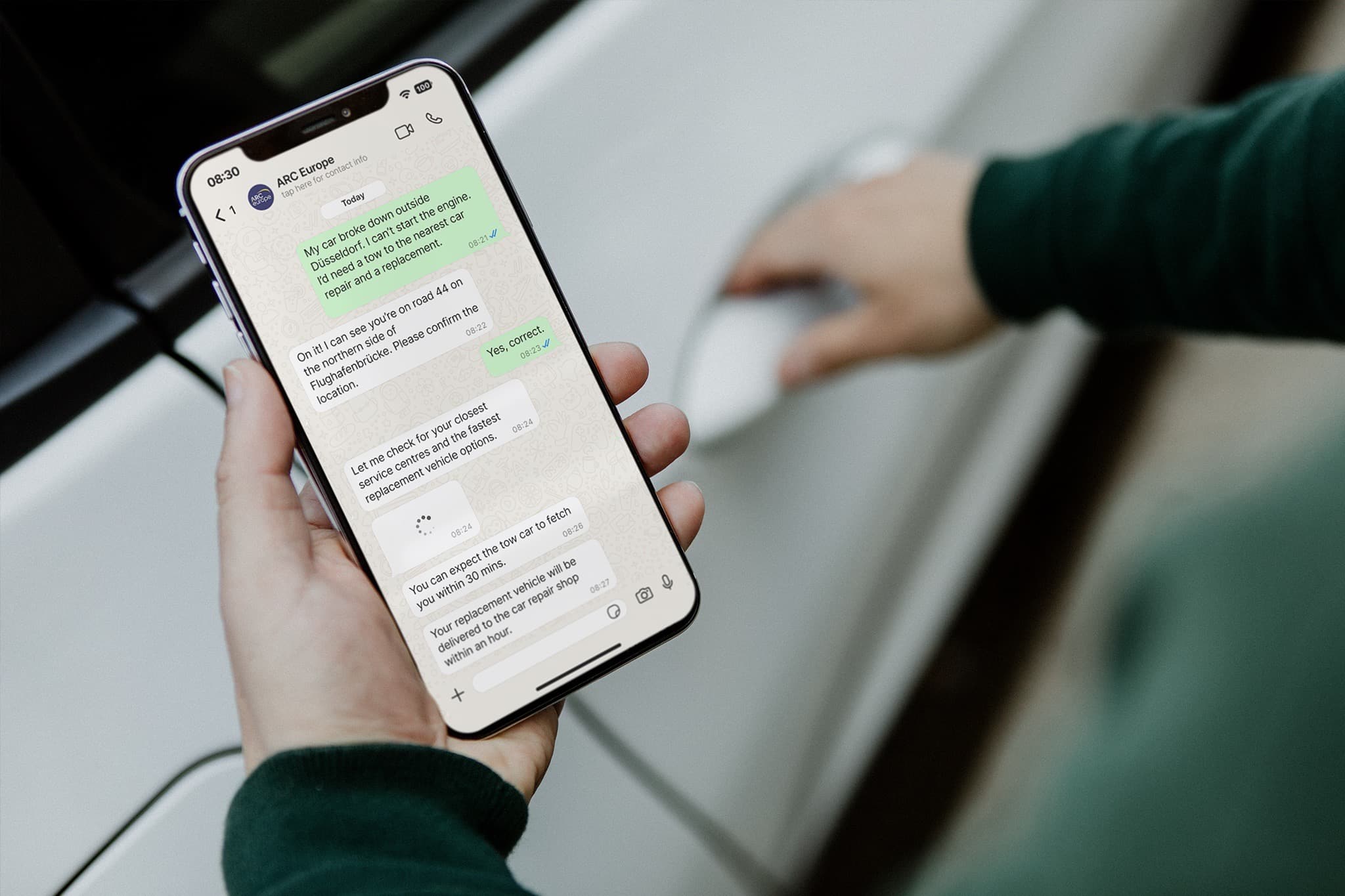 Driver holding a smartphone showing the WhatsApp assistance chatbot next to a car