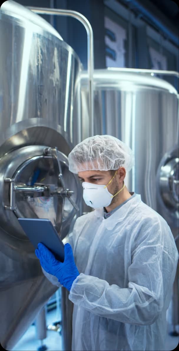 Technician in protective clothing using a tablet beside stainless steel tanks