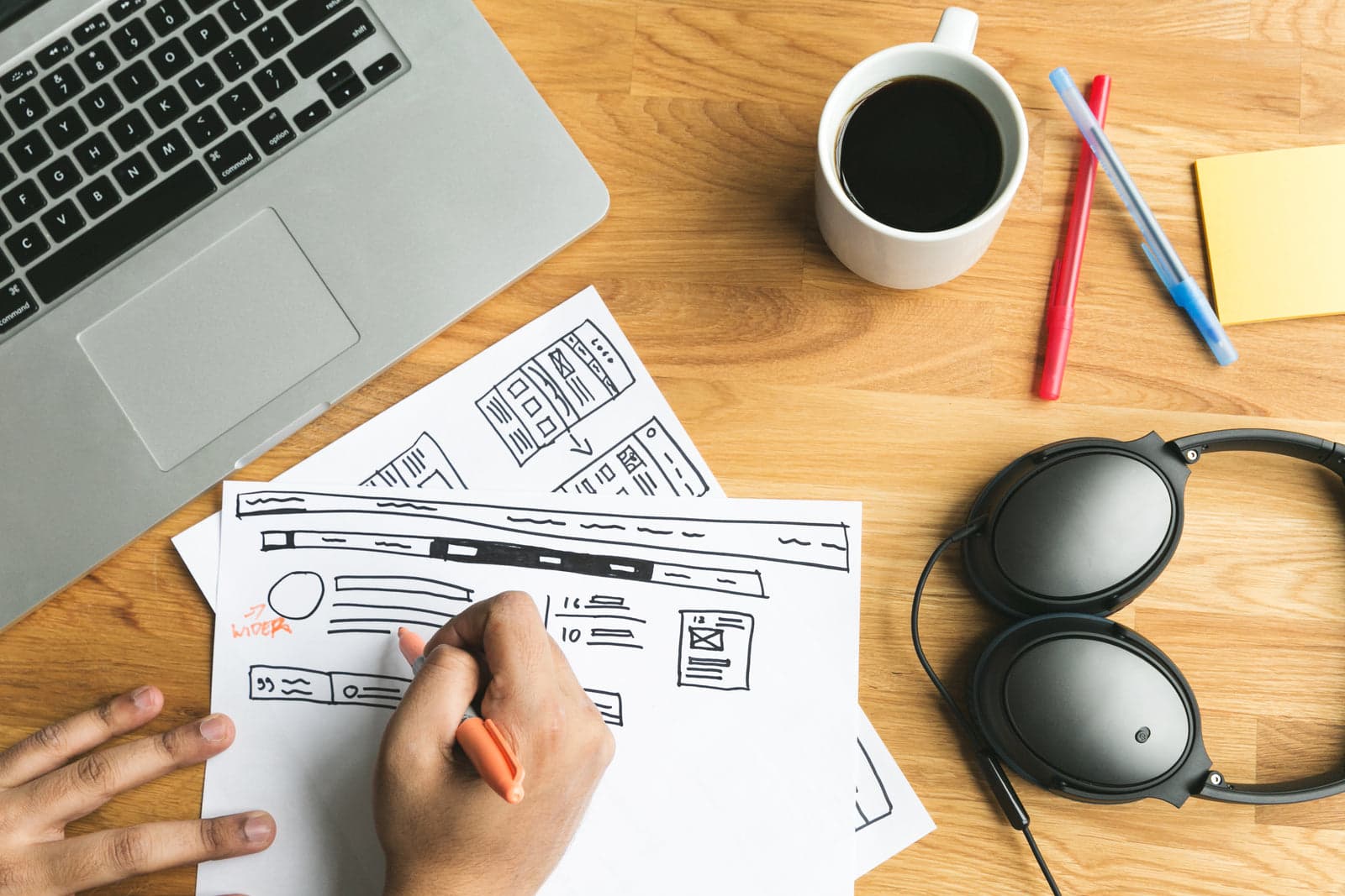 hands sketching UI wireframes next to a laptop